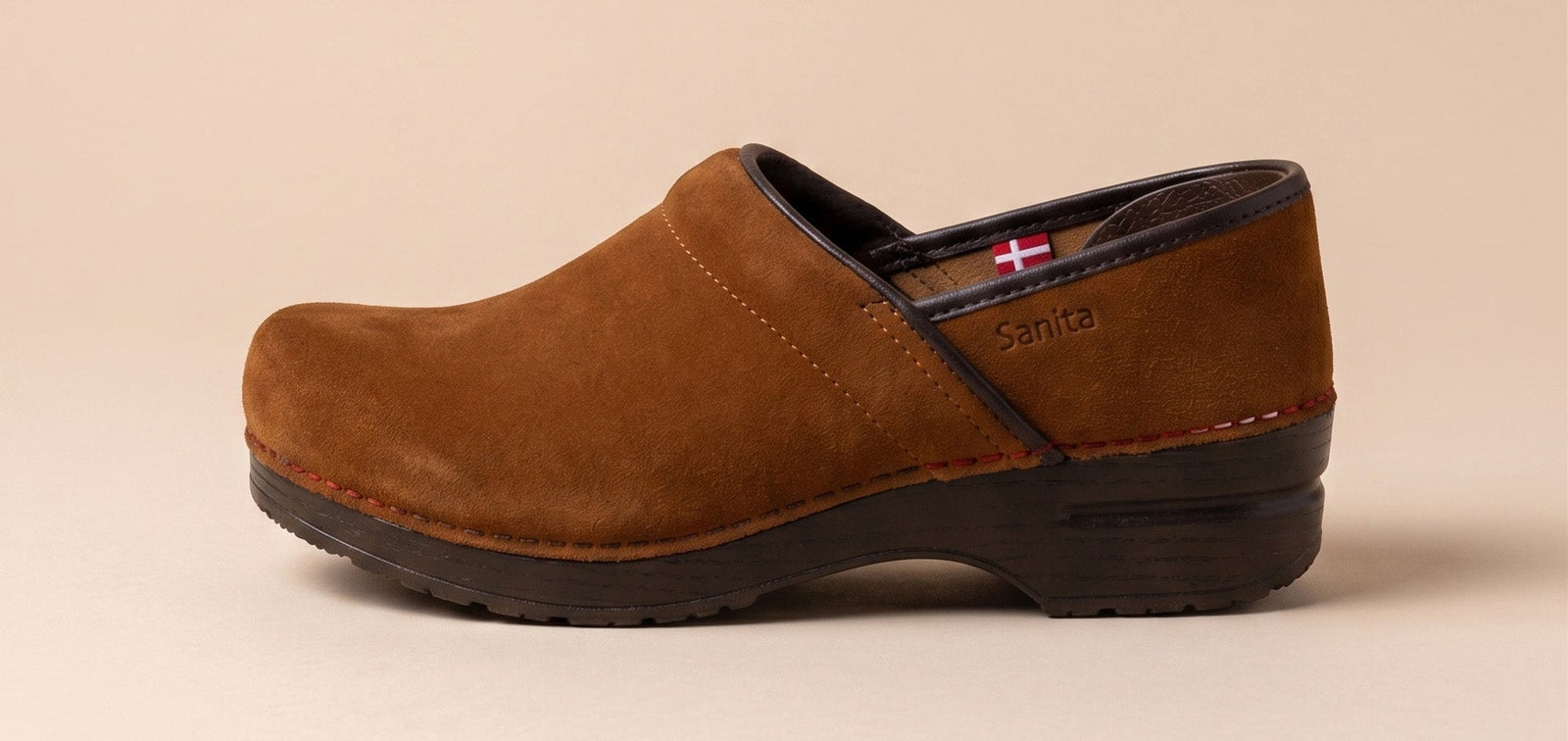 A single brown suede clog shoe shown in profile against a solid beige background. The shoe features a dark brown sole and heel counter with a small red and white flag detail on the inner lining.