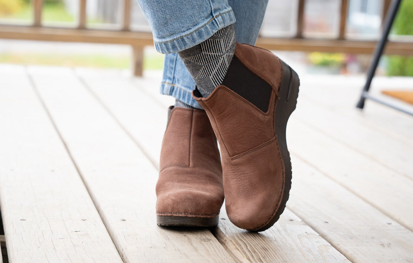 A person wearing brown suede ankle boots with black elastic side panels, paired with light blue jeans, standing on a light-colored wooden deck.